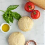 2 balls of whole wheat pizza dough on a white board with tomatoes, basil, and olive oil.
