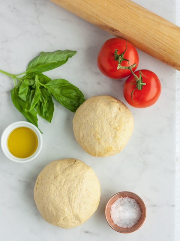 2 balls of whole wheat pizza dough on a white board with tomatoes, basil, and olive oil.