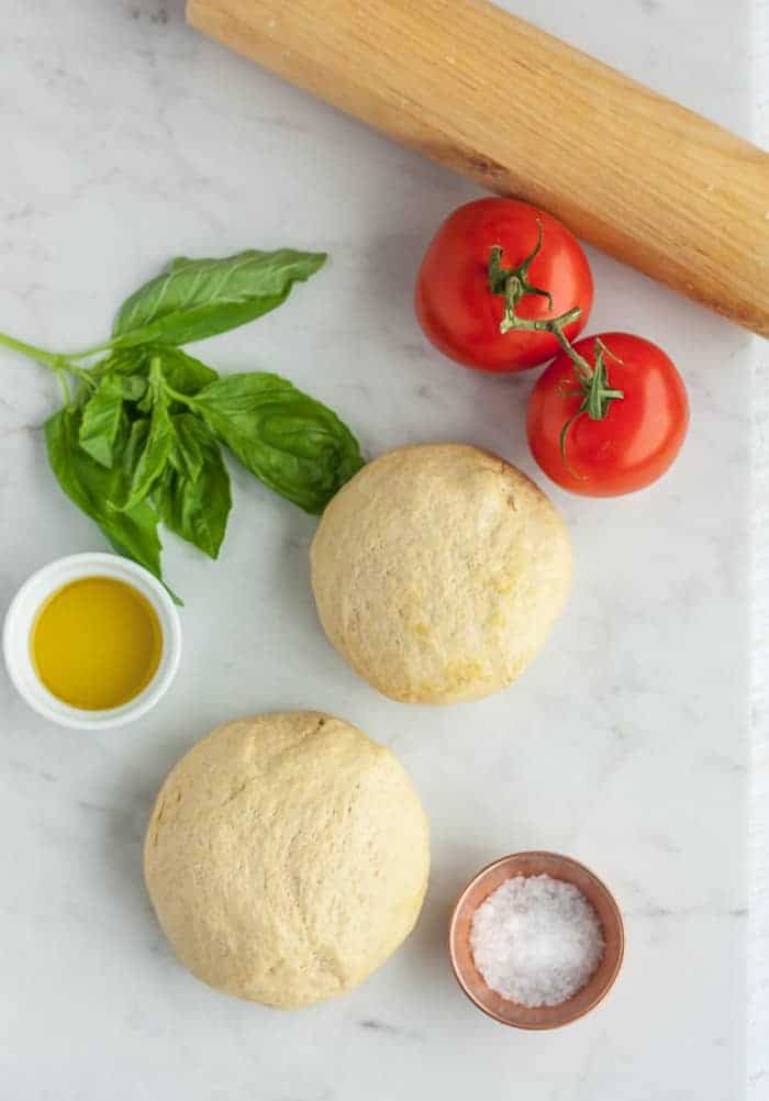 2 balls of whole wheat pizza dough on a white board with tomatoes, basil, and olive oil.