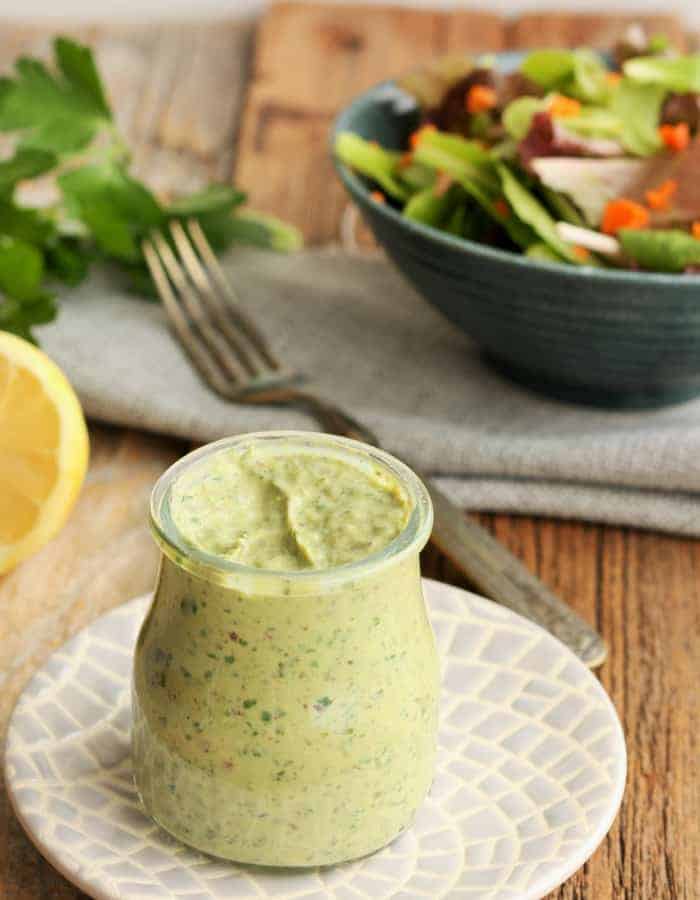 A container of green goddess dressing on a plate with a salad and fork in the background