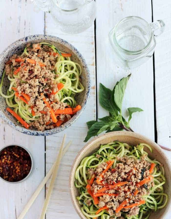 Two bowls of a thai basil chicken recipe, basil, glasses, and chopsticks on a white board