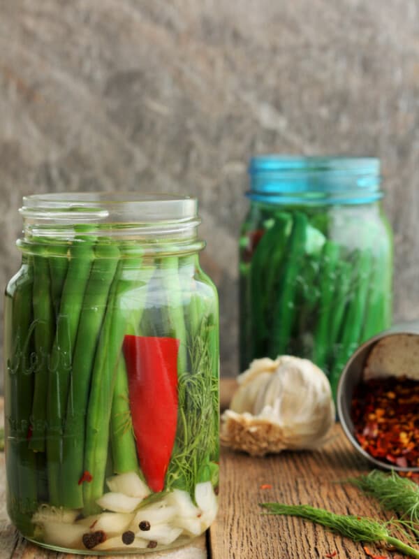 Two jars of dilly beans with garlic and red peppers on a wooden board