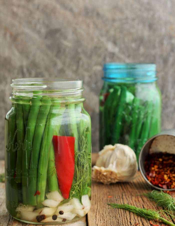 Two jars of dilly beans with garlic and red peppers on a wooden board