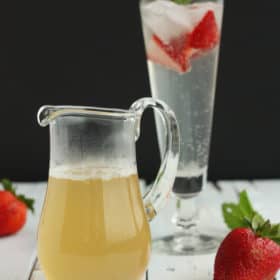 A small glass pitcher of honey simple syrup in front of a glass with strawberries, mint, and sparkling water