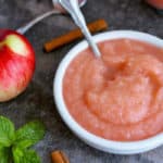 A bowl of bowl of homemade applesauce with cinnamon sticks and apples on a grey board