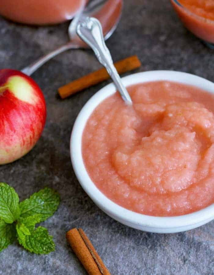 A bowl of bowl of homemade applesauce with cinnamon sticks and apples on a grey board