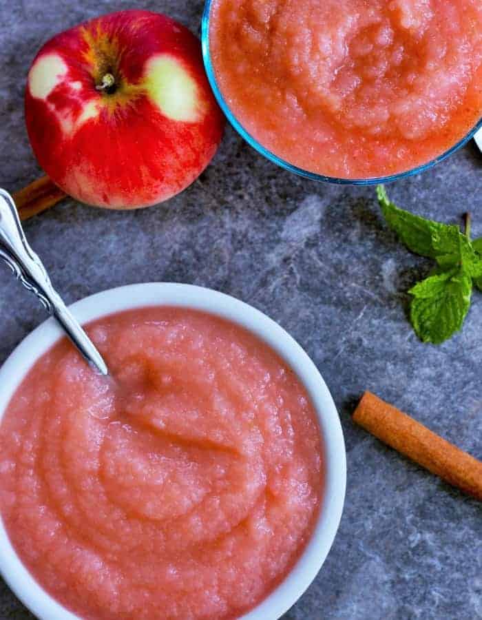 Bowls of homemade applesauce with a cinnamon stick and apples on a grey board