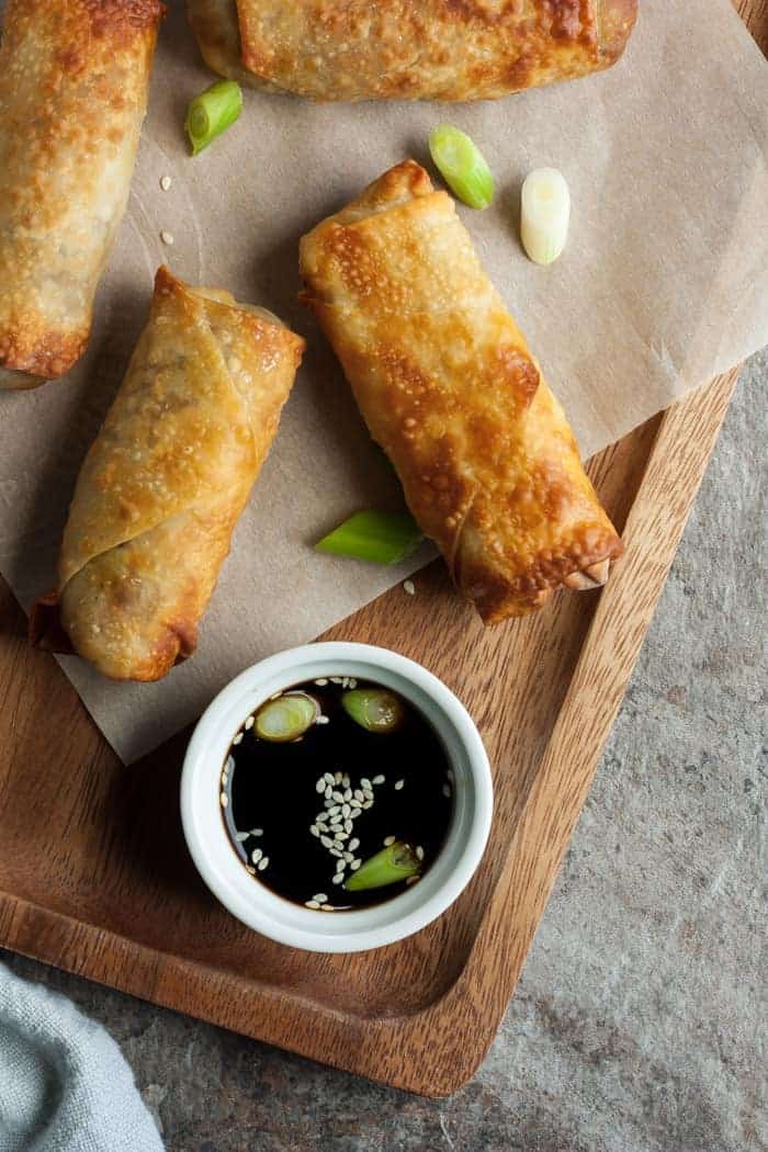 Three air fryer egg rolls on a wooden tray with a dish of soy sauce.