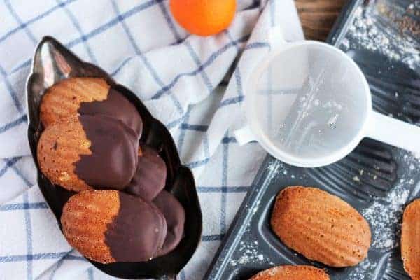 chocolate dipped madeleines on a tray with an orange and sieve on a madeleine tray