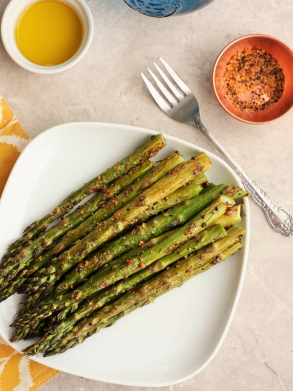 A plate of roasted asparagus on a tray with a fork and seasoning