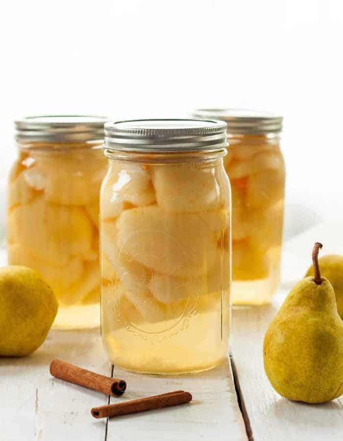 Three jars of canned pears on a wooden board with fresh pears and cinnamon sticks