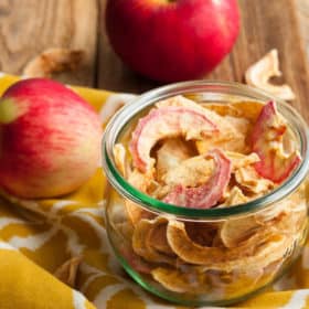 A glass jar of dried apples with a yellow cloth and apples on a wooden board