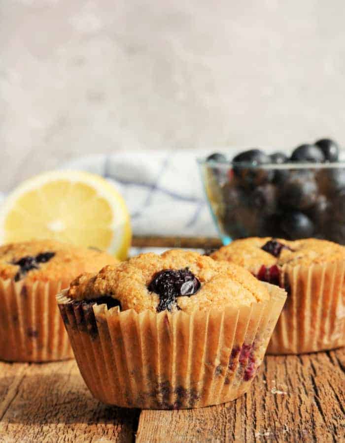 Three whole wheat blueberry muffins on a wooden board with a lemon and a bowl of blueberries