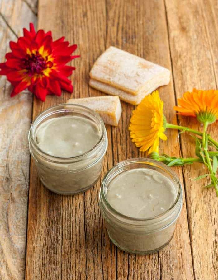 two jars of a homemade deodorant recipe with flowers and beeswax on a wooden board