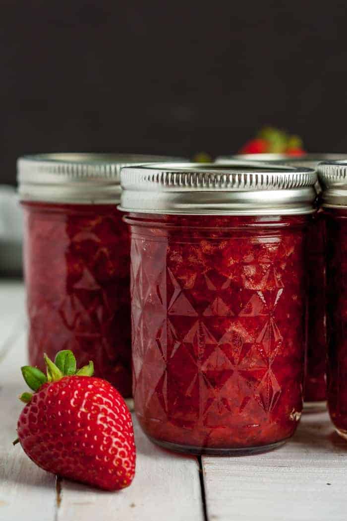Three jars of homemade strawberry jam with fresh strawberries on a white board