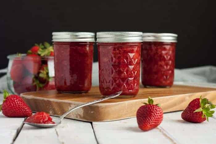 Three jars of homemade strawberry jam on a cutting board with strawberries and a spoon