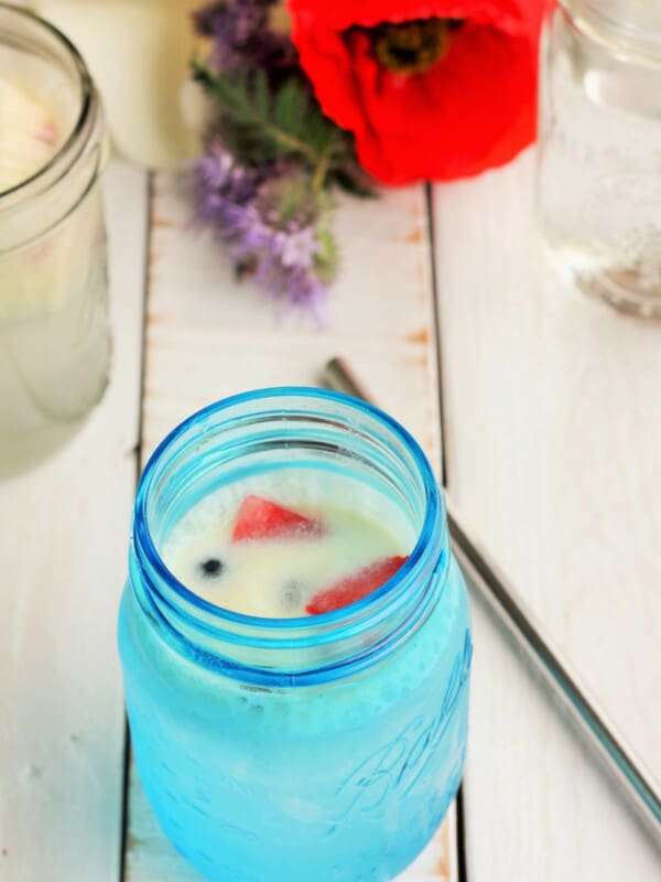 a blue mason jar with creamy berry part drink, a straw, and flowers on a white board