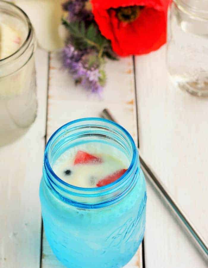 a blue mason jar with creamy berry part drink, a straw, and flowers on a white board