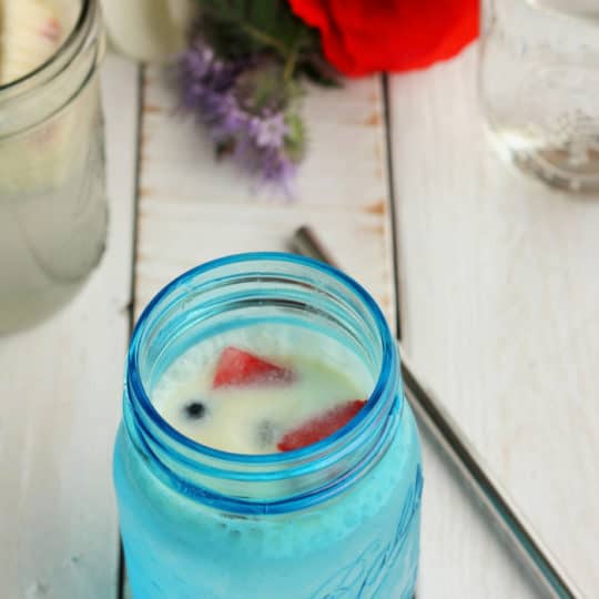 blue mason jar with a creamy berry drink and straw on a white board with flowers