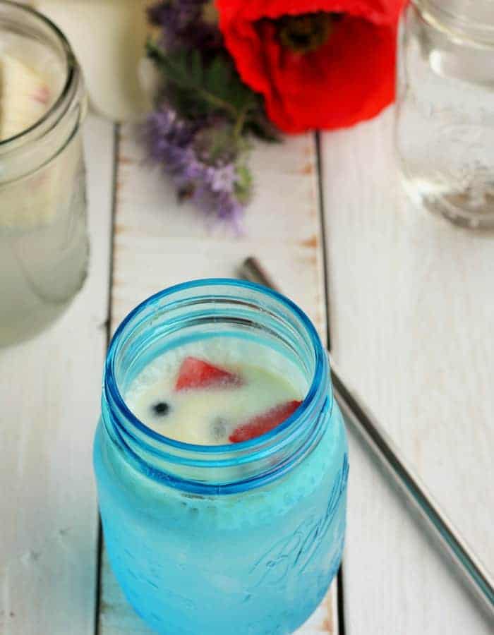 blue mason jar with a creamy berry drink and straw on a white board with flowers