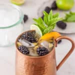 a peach moscow mule with blackberries and mint on a white board