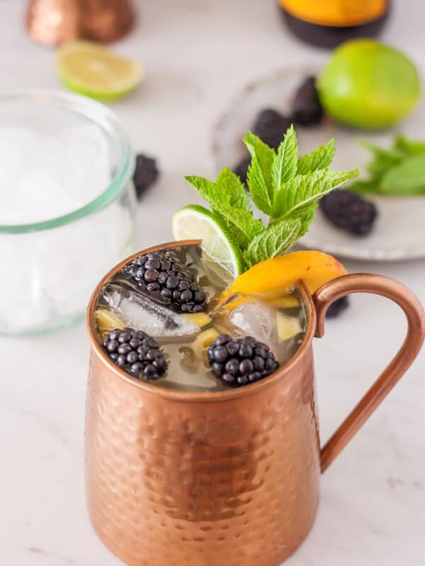 a peach moscow mule with blackberries and mint on a white board