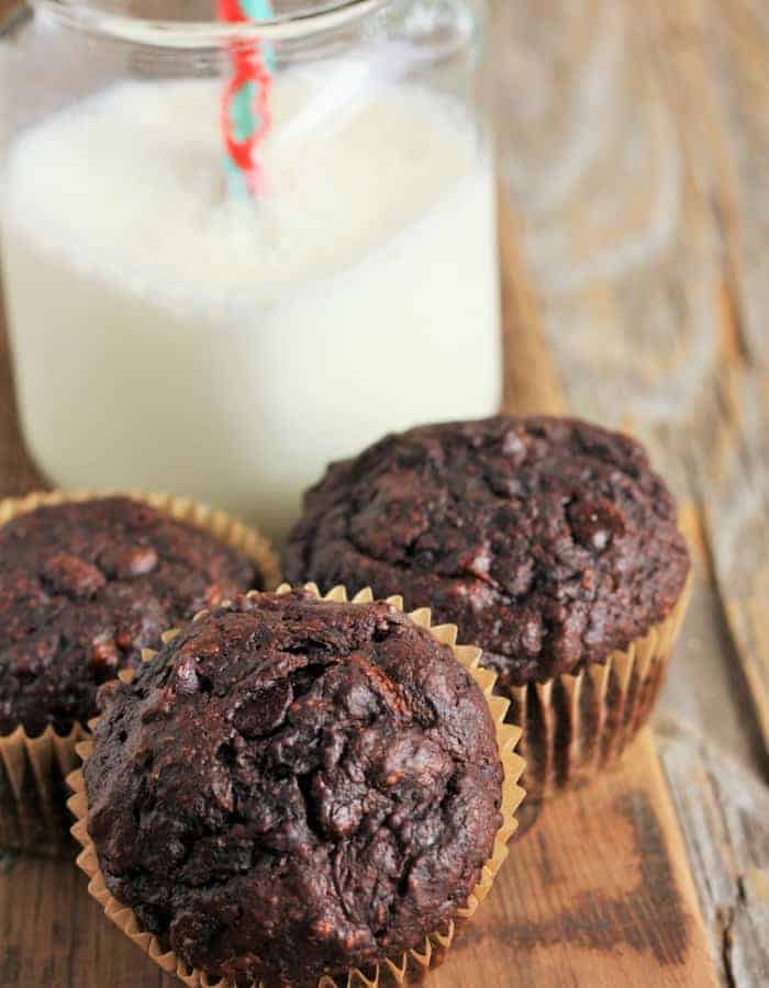 three double-chocolate zucchini muffins and a glass of milk with a straw on a wooden board