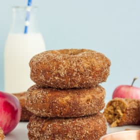 3 cinnamon apple donuts stacked up with apples and a bottle of milk