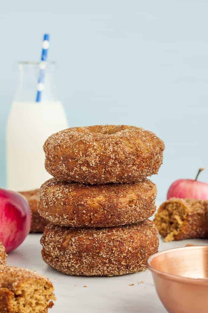 3 cinnamon apple donuts stacked up with apples and a bottle of milk