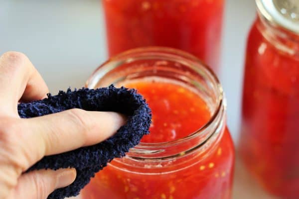 a washrag cleaning the rim of a canning jar full of tomatoes