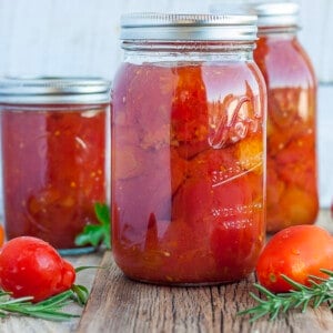 three jars of whole peeled tomatoes with fresh tomatoes and herbs on a wooden board