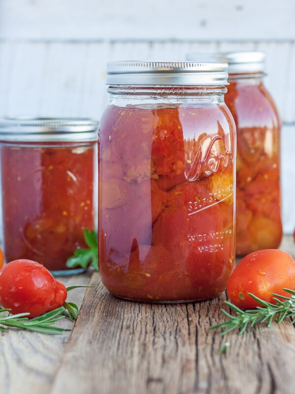 three jars of whole peeled tomatoes with fresh tomatoes and herbs on a wooden board