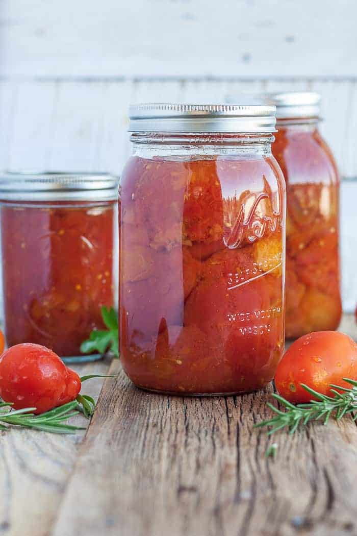 three jars of whole peeled tomatoes with fresh tomatoes and herbs on a wooden board