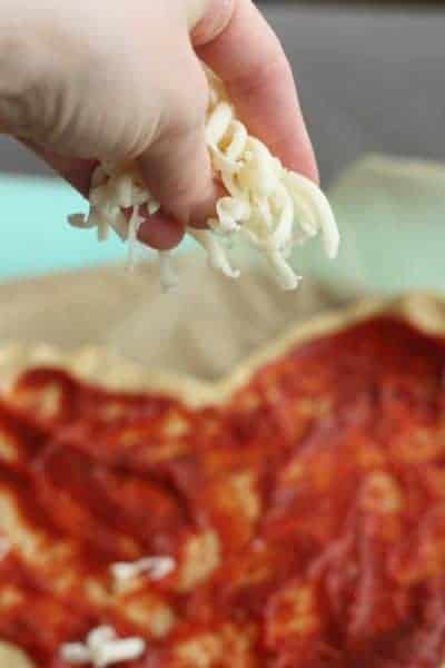 A hand holding shredded mozzarella cheese over a heart-shaped pizza | www.sustainablecooks.com