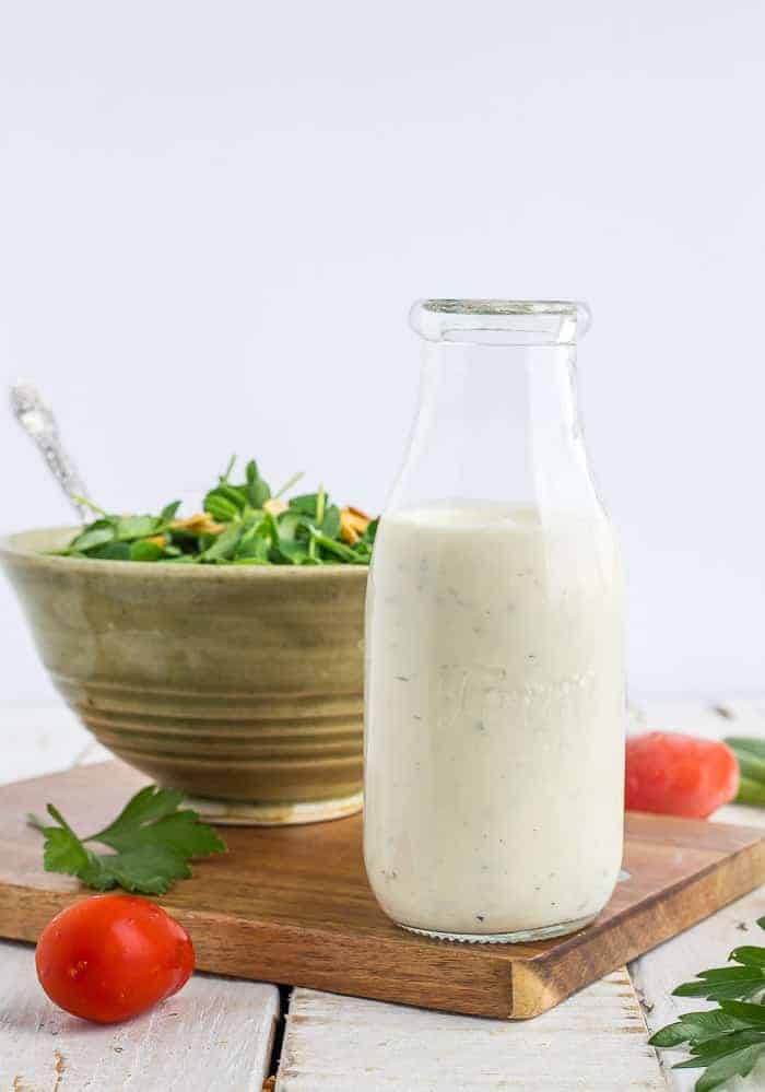 A glass bottle of ranch dressing in front of a bowl of salad