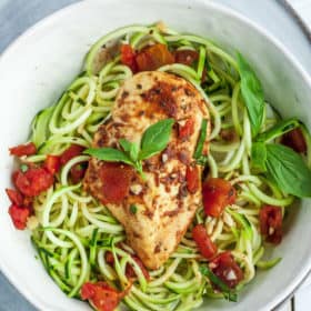 a white bowl with zoodles and bruschetta chicken topped with basil and tomatoes