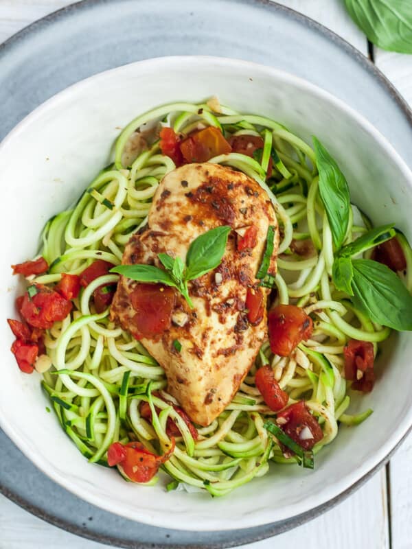 a white bowl with zoodles and bruschetta chicken topped with basil and tomatoes