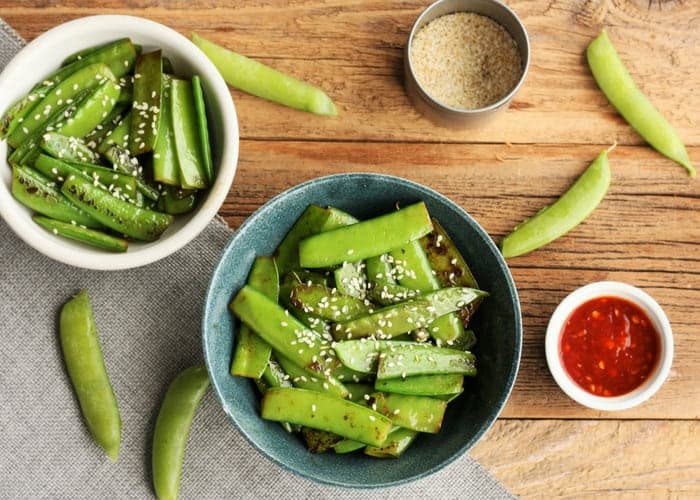 Two bowls of sesame snow peas with chili paste, sesame seeds, and pea pods on a wooden board