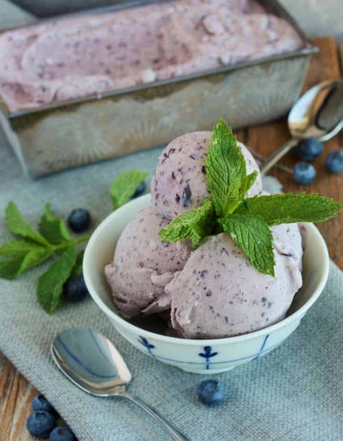 A bowl of Fresh blueberry ice cream with mint, spoons, and blueberries on a wooden board