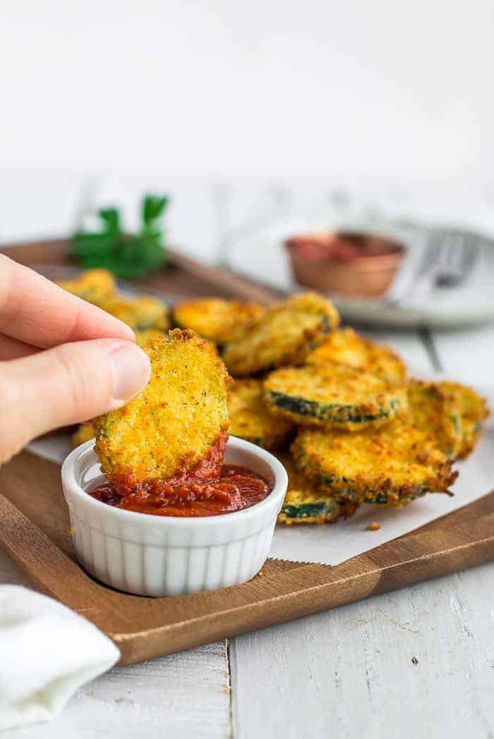 a hand dipping fried zucchini in a white bowl of marinara