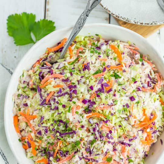 a multi-colored coleslaw in a white bowl with a spoon on a white board