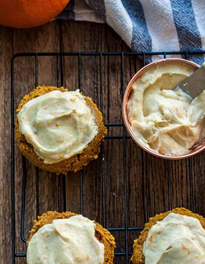 Nothing welcomes Fall like baking up a batch of Grandma Dorothy's Soft Pumpkin Cookies for dessert! These amazing cookies are so easy to make and are topped with an incredible orange cream cheese frosting. #sustainablecooks #pumpkincookies #wholewheatbaking #pumpkindesserts frosted soft pumpkin cookies on a baking rack with a dish of frosting and a knife