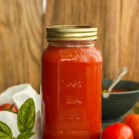 A jar of home canned tomato soup with a bowl of soup and herbs and tomatoes