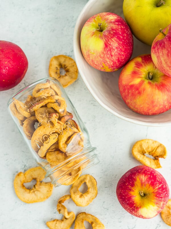 a jar of dried apples tipped on its side with a bowl of apples on a white board