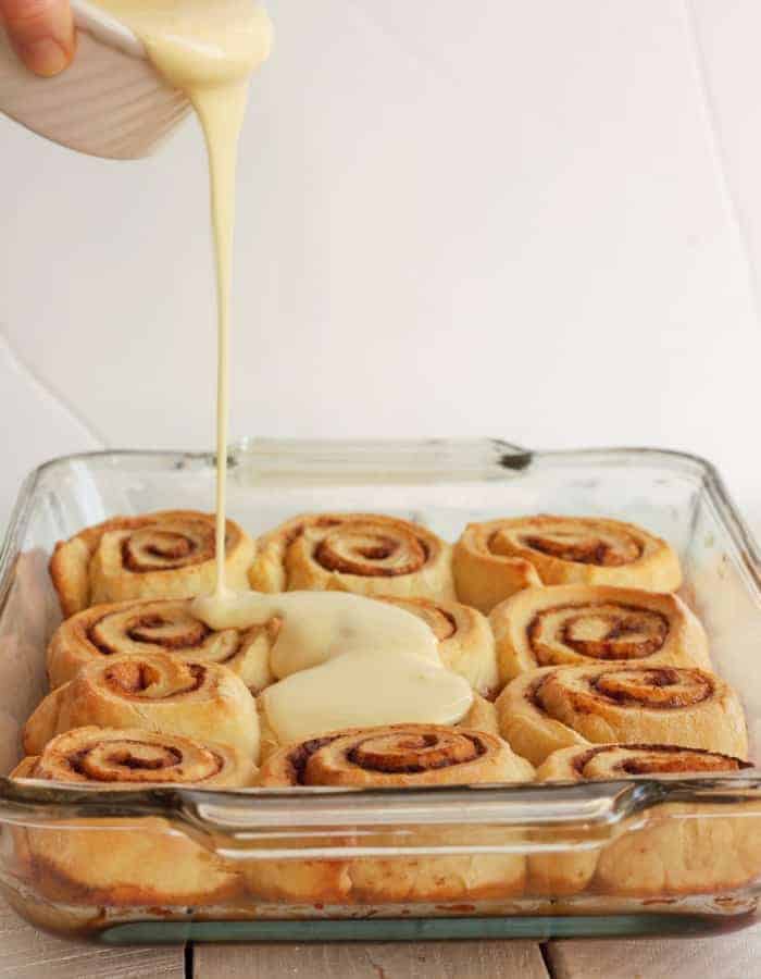 A bowl of orange cream cheese frosting being poured over overnight cinnamon rolls