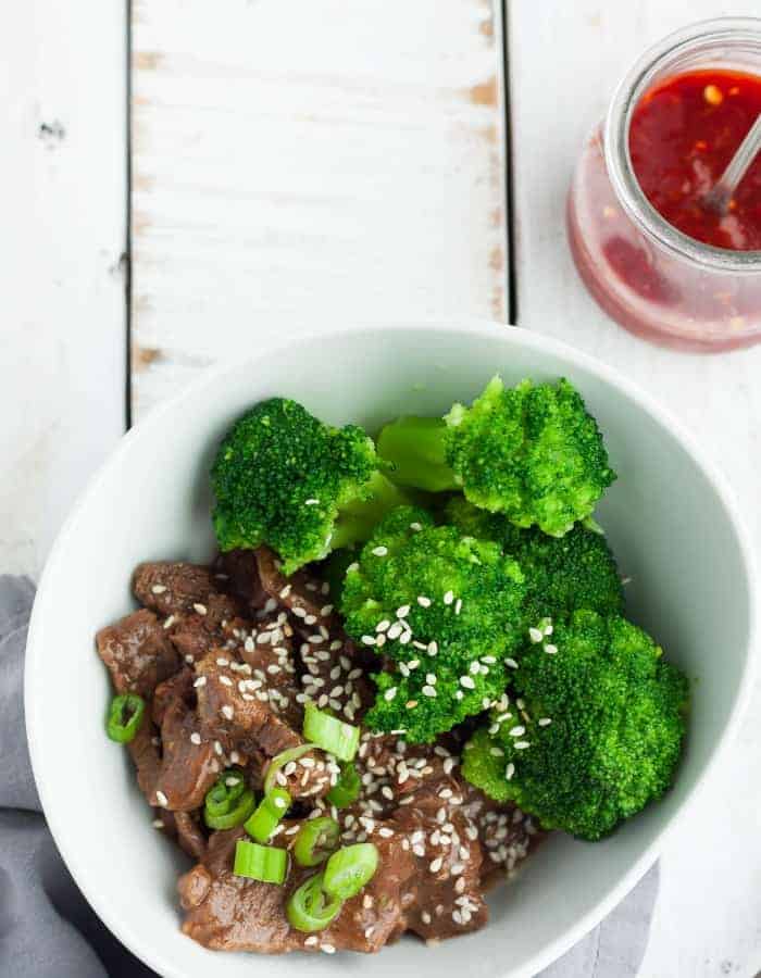 An overhead photo of a bowl of paleo beef and broccoli with sesame seeds and hot sauce.