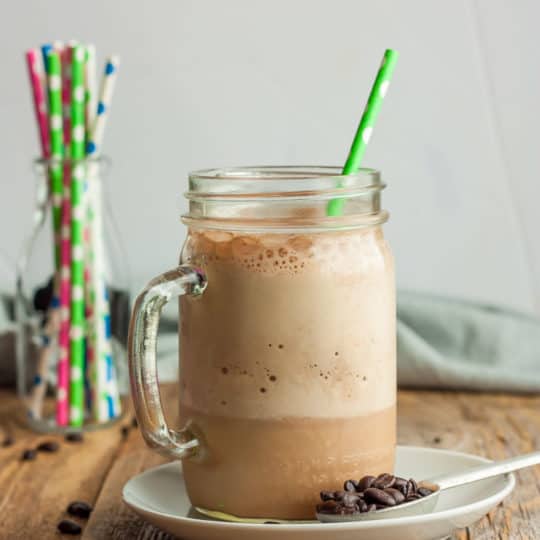 a canning jar glass of blended iced coffee with a green straw on a plate with a spoon and coffee beans.