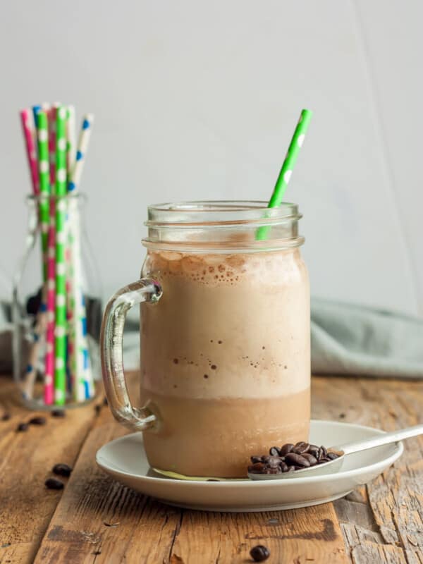 a canning jar glass of blended iced coffee with a green straw on a plate with a spoon and coffee beans.