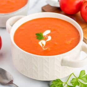 a white bowl of dairy-free tomato soup on a white board with tomatoes and fresh parsley