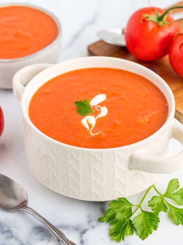 a white bowl of dairy-free tomato soup on a white board with tomatoes and fresh parsley